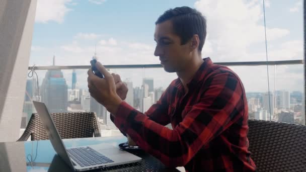 Busy business man uses smartphone, works at a laptop and drinks coffee in a cafe on the high floor of a skyscraper on background of the big city of the metropolis. Remote work, freelance concept — Wideo stockowe