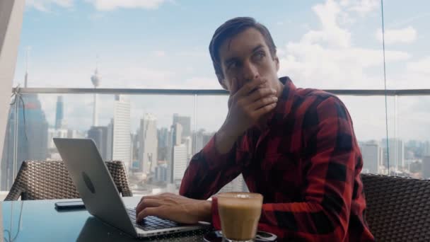 Man is unpleasantly surprised and shocked by the bad news seen on the laptop screen. Man suffers in stress and despair, while sitting at cafe on background of Big City Skyscrapers. — Stok video