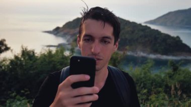 Man Tourist with Backpack Using Smartphone. Traveler on top of a mountain on the background of a tropical island in the sea. Travel concept