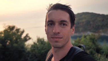 Portrait of man tourist making selfie or a video call using smartphone outdoors on background of evening tropical island sunset