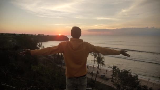 Young man traveler raising his hands high on top of the big mountain above the ocean and beautiful tropical beach on golden sunset — 비디오