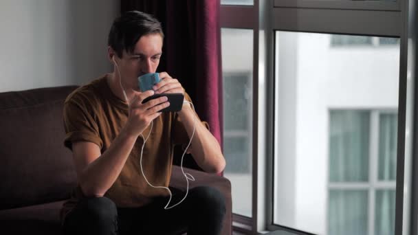 Homme souriant dans les écouteurs bénéficie d'un smartphone regarder les médias dans les réseaux sociaux tout en étant assis à la maison sur le canapé et boit un délicieux café . — Video