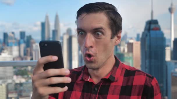 Happy Man in Plaid Shirt Celebrating Win on Smartphone on the roof of skyscraper. Emotions of joy and delight. Lottery win concept. Background of the big city skyscrapers. — Stockvideo