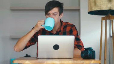 Genç erkek fotoğrafçı dizüstü bilgisayarında resim işliyor ve evde kahve içiyor..