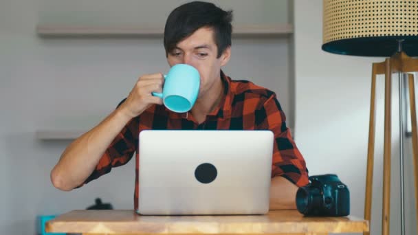 Junger männlicher Fotograf mit Laptop, der Bilder auf seinem Laptop bearbeitet und zu Hause Kaffee trinkt. — Stockvideo