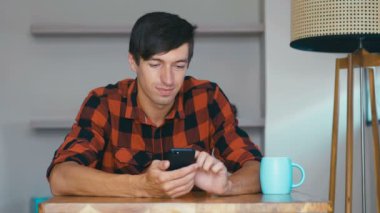 Neşeli genç adam akıllı telefon kullanıyor evde gülümseyerek eğleniyor evde tek başına dinleniyor.