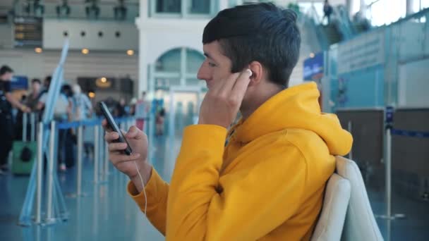 Giovane seduto all'aeroporto vicino alla finestra e utilizza il telefono, aeroplano offuscato sullo sfondo. Guy in attesa di volo nella lounge dell'aeroporto — Video Stock