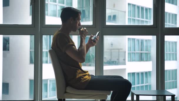 Молодой человек сидит перед окном, пьет кофе и по утрам серфингует смартфон дома — стоковое видео