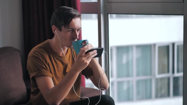 Jovem em fones de ouvido usando smartphone assistindo mídia em redes sociais enquanto sentado em casa no sofá e bebe delicioso café . — Vídeo de Stock
