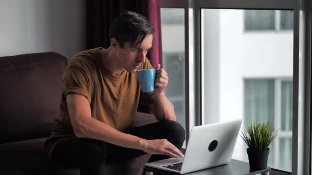 Hombre bebiendo café mientras usa un ordenador portátil en casa sentado en el sofá — Vídeo de stock