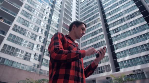 Bonito jovem em camisa xadrez navegando seu Smartphone na cidade em prédios urbanos fundo . — Vídeo de Stock