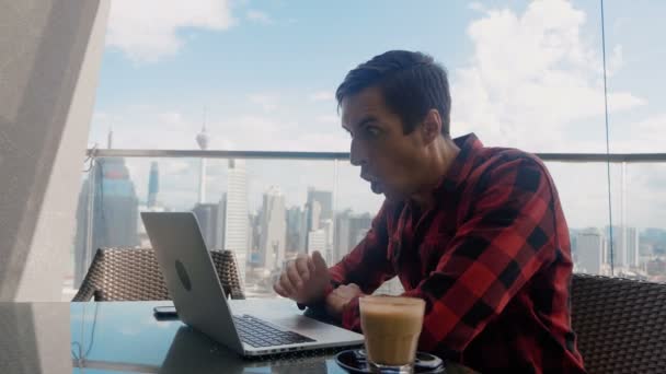 Man is unpleasantly surprised and shocked by the bad news seen on the laptop screen. Man suffers in stress and despair, while sitting at cafe on background of Big City Skyscrapers. — Wideo stockowe