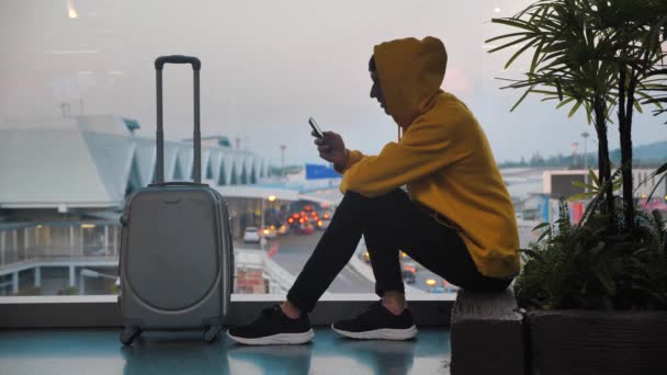 Silhouet van hooded man toerist met bagage te wachten op de luchthaven terminal zit in de buurt venster, reiziger met behulp van de smartphone en wachten op instappen. Vliegtuig op achtergrond. — Stockvideo