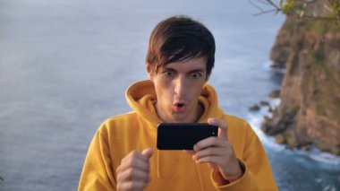 Deniz dalgalarının üzerindeki bir uçurumda akıllı telefon uygulamasında kazanan bir erkek gezgin. Kutlama kazananı iyi haber ya da online oyun uygulamasında seyahat ederken başarı..