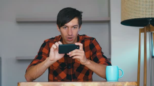 Akıllı telefonlu oyun oynayan genç adam evde bir masaya otur, sosyal medya uygulamasını kullanan mutlu genç adam ekrandaki telefona bakarak cep telefonu çalışmasını izle. — Stok video