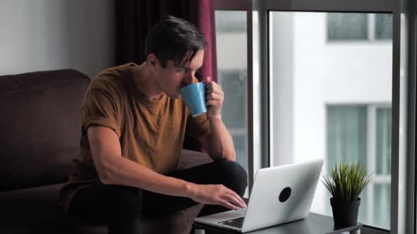 Knappe jongeman drinken koffie tijdens het gebruik van een laptop computer, thuis zitten op een bank voor een raam. — Stockvideo