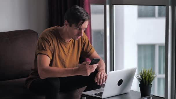 Joven ocupado usando el teléfono inteligente y el ordenador portátil para el comercio electrónico haciendo compras en línea viendo la película de vídeo sentado en el sofá en casa — Vídeo de stock