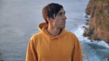 Close up portrait of young man traveler looking at the camera on the top of the mountain, on background of cliff and ocean waves