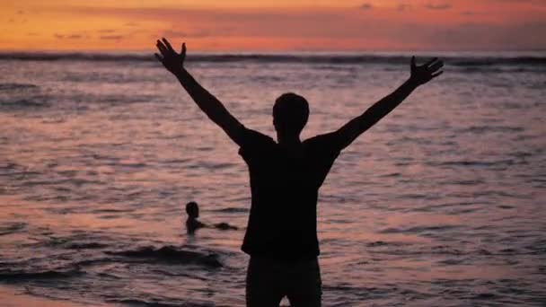 男のシルエットは、赤い夕日の美しい熱帯のビーチに立って手を上げる。自由の概念. — ストック動画