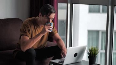 Handsome young man uses a laptop computer and drinks coffee in the morning while sitting at home on the sofa in front of the window.