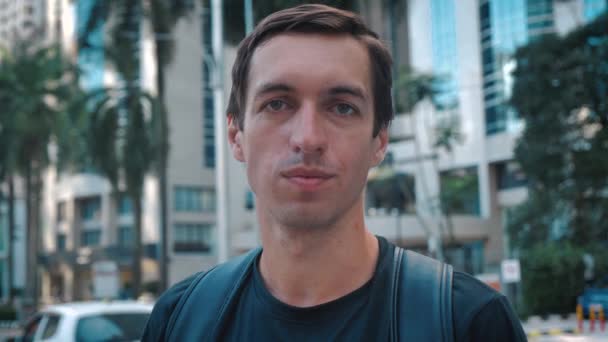 Retrato de un joven guapo turista con mochila parado al aire libre solo sonriendo mirando a la cámara en el fondo ciry urbano — Vídeo de stock