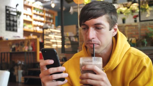 Человек, использующий сенсорный экран смартфона в современном кафе. Мужчина пьет молочный коктейль или смузи и смс-ки на смартфоне в кафе — стоковое видео