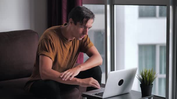 Jovem bem sucedido usando laptop fica surpreso, então feliz e comemora vitória enquanto sentado no sofá na sala de estar em casa contra a janela — Vídeo de Stock