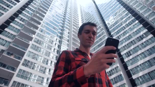 Retrato de un joven con camisa a cuadros navegando por su Smartphone en la ciudad. Guapo hombre seguro de comunicarse en el teléfono inteligente en los edificios urbanos de fondo . — Vídeo de stock