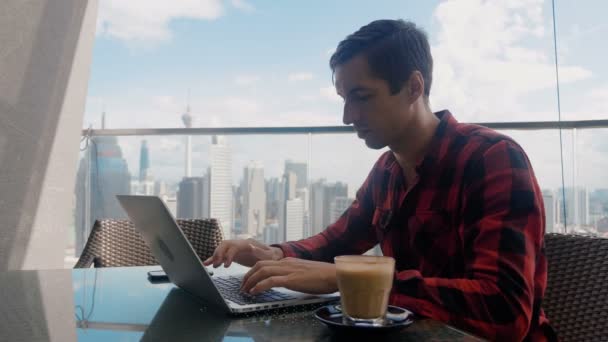 Homem de negócios confiante usando smartphone e laptop no café no telhado de um arranha-céu contra o fundo de um arranha-céus grande metrópole da cidade . — Vídeo de Stock