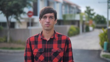 Portrait of handsome caucasian man in red plaid shirt standing in front of the camera and smiling at suburb of the city on the background of houses