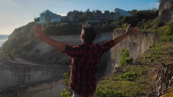 Young man traveler raising his hands high on top of the mountain above beautiful landscape on golden sunset — Stockvideo