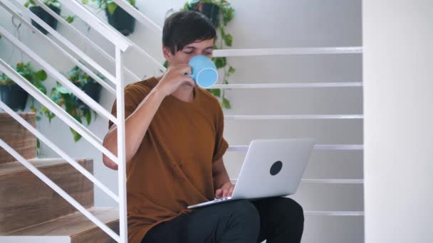 Молодий чоловік-фрілансер, який використовує ноутбук, який вивчає онлайн, працюючи з дому в Інтернеті, посміхається тисячолітній хлопець, друкує на комп'ютері і п'є каву, сидячи на сходах вдома . — стокове відео