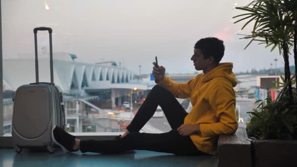 Joven sonriente Turista con equipaje esperando en la terminal del aeropuerto pecando ventana ordenada, viajero mensajes de texto en el teléfono inteligente y esperando a embarque — Vídeo de stock