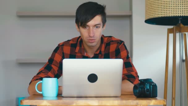 Όμορφος φωτογράφος με κάμερα και φορητό υπολογιστή κάθεται στο τραπέζι στο σπίτι, πίνοντας καφέ και επεξεργάζεται φωτογραφίες με retouch — Αρχείο Βίντεο