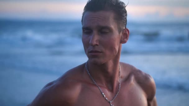 Retrato del hombre atlético guapo, el deportista musculoso bien entrenado, posando en la playa al atardecer hermoso — Vídeo de stock