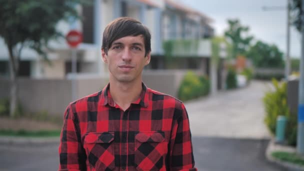 Portrait of handsome caucasian man in red plaid shirt standing in front of the camera and smiling at suburb of the city on the background of houses — 图库视频影像