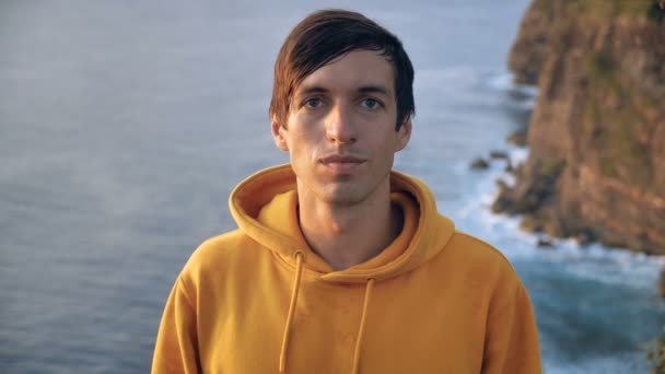 Close up portrait of young man traveler looking at the camera on the top of the mountain, on background of cliff and ocean waves — Stok video