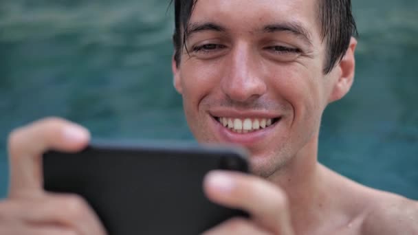 Primer plano del joven sonriente de vacaciones viendo vídeo en un smartphone en una piscina — Vídeos de Stock