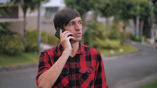 Портрет красивого кавказца в красной клетчатой рубашке, разговаривающего по сотовому телефону в пригороде города на фоне домов — стоковое видео