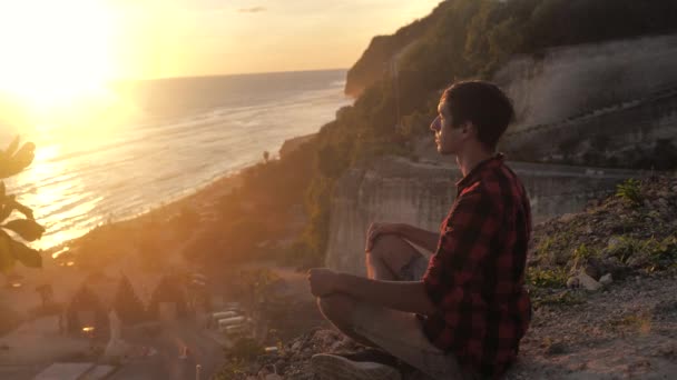 Az ember meditál, miközben ül egy sziklán szemben egy gyönyörű naplemente. — Stock videók