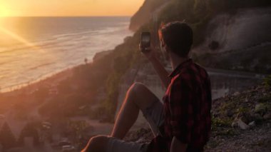 Genç adamın silueti akıllı telefonuyla güzel günbatımının fotoğrafını ya da videosunu çeker. Seyahat blogcusu sosyal medya için fotoğraf yapıyor.