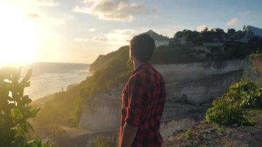 Genç adam gün batımının tadını çıkarırken denizin arka planında bir kayanın üzerinde duruyor.