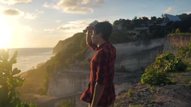 Genç adam gün batımının tadını çıkarırken denizin arka planında bir kayanın üzerinde duruyor.