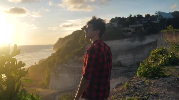 Joven viajero masculino disfrutando de una hermosa puesta de sol mientras está de pie sobre una roca sobre un fondo del mar — Vídeos de Stock