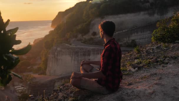 Homem medita enquanto sentado em uma rocha contra um belo pôr do sol . — Vídeo de Stock