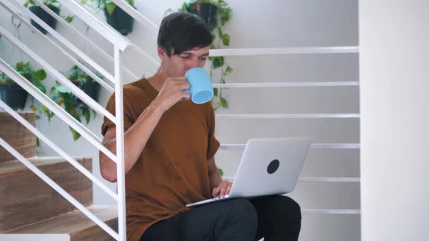 Jeune homme utilisant un ordinateur portable et boire du café tout en étant assis sur les escaliers à la maison . — Video