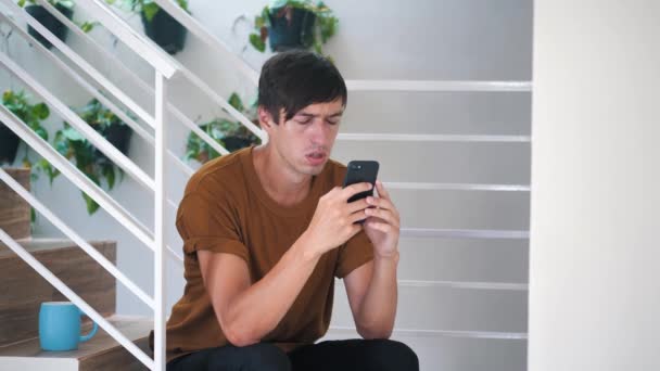 Smutný mladý muž sedí doma na schodech a používá smartphone. — Stock video