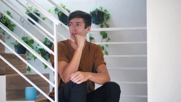 Pensive sad and depressed young man sitting alone on stairs at home: painful sad man. — ストック動画