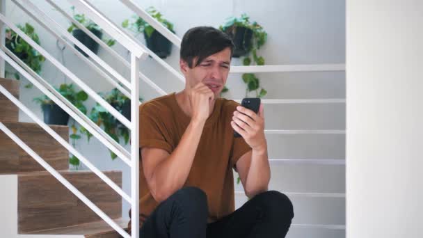 Chockad Ung Man Reagerar på Förlust på meddelande på Smartphone. Obehagligt överraskad och rädd. Sad Man fick dåligt meddelande på sin smartphone, sitter på trappor hemma. — Stockvideo