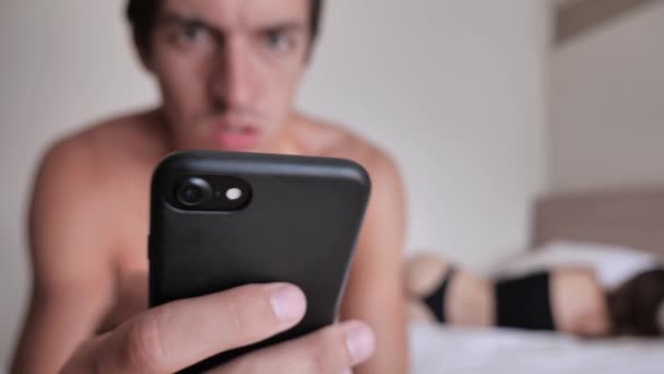 Een stel heeft problemen in bed. Bezorgd Droevige man gebruikt een smartphone in de slaapkamer zittend op bed. Seksuele problemen, problemen in relaties — Stockvideo